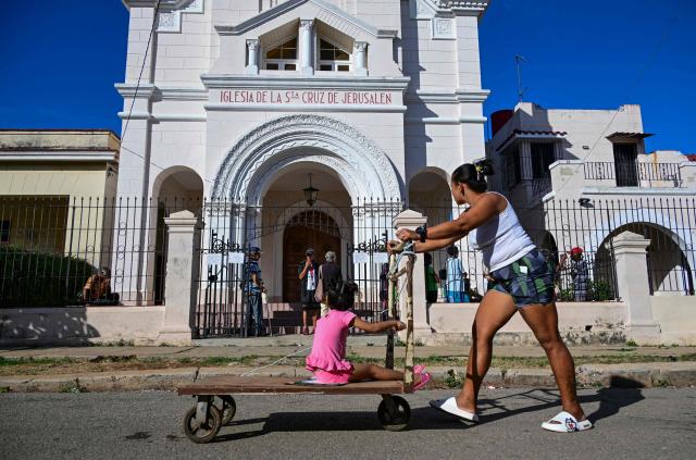 A woman pushes a cart with her daughter past the Church of the Holy Cross of Jerusalem in Havana on April 14, 2026. Dozens of people, mainly elderly and low income persons, come to this church to receive free medicines they cannot find in pharmacies or hospitals, reflecting the social role churches are playing in a Cuba in crisis. (Photo by YAMIL LAGE / AFP)