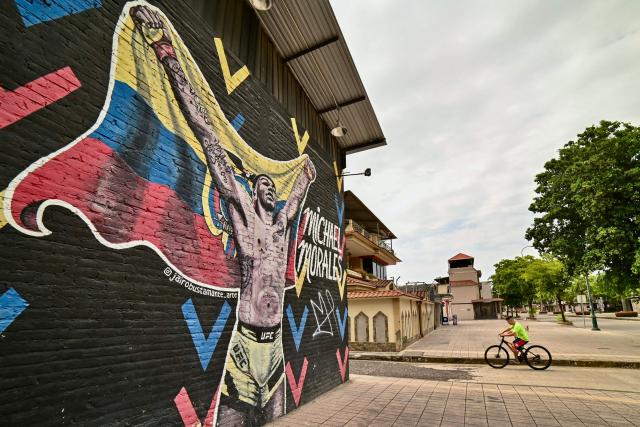 A man rides a bike past a mural depicting Ecuadorian mixed martial arts fighter Michael Morales outside his former training gym in Machala, El Oro province, Ecuador on April 4, 2026. In a tropical town in Ecuador, children no longer dream only of becoming football players to escape poverty. They also want to be UFC fighters like Michael "Spiderman" Morales, a beast in the octagon who has become a local idol. The 26-year-old fighter has been immortalized in murals and is now the hero of the children on the streets of Pasaje, a town ravaged by violence. (Photo by Marcos PIN / AFP)
