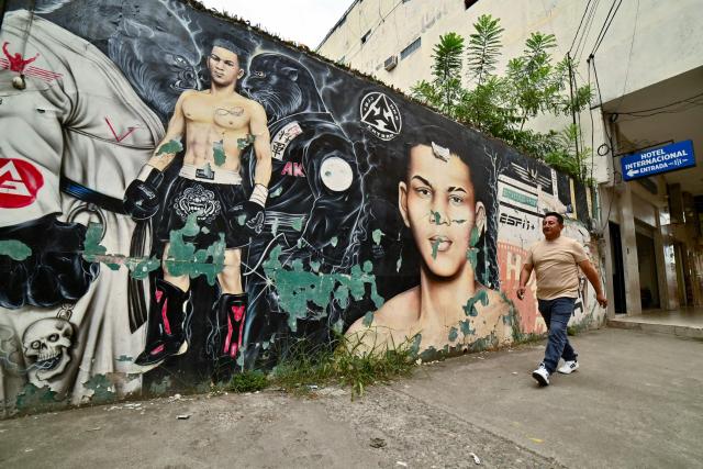 A man walks past a mural depicting Ecuadorian mixed martial arts fighter Michael Morales in Pasaje, El Oro province, Ecuador on April 4, 2026. In a tropical town in Ecuador, children no longer dream only of becoming football players to escape poverty. They also want to be UFC fighters like Michael "Spiderman" Morales, a beast in the octagon who has become a local idol. The 26-year-old fighter has been immortalized in murals and is now the hero of the children on the streets of Pasaje, a town ravaged by violence. (Photo by Marcos PIN / AFP)