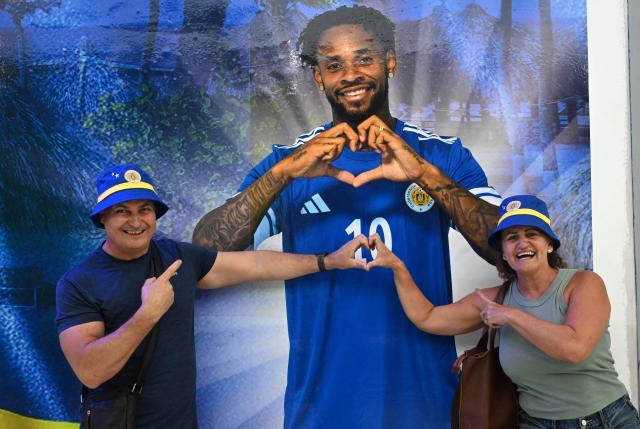 A couple makes a heart next to an image of Curacao's player Leandro Bacuna at the official fan store in Willemstad, Curacao, in the Dutch Caribbean, on April 8, 2026. Small island, big dreams. The poster of Curacao’s national football team hangs at the entrance of Marchena, a working-class neighborhood of Willemstad, the capital of the smallest territory ever to qualify for a World Cup. (Photo by Raul ARBOLEDA / AFP)