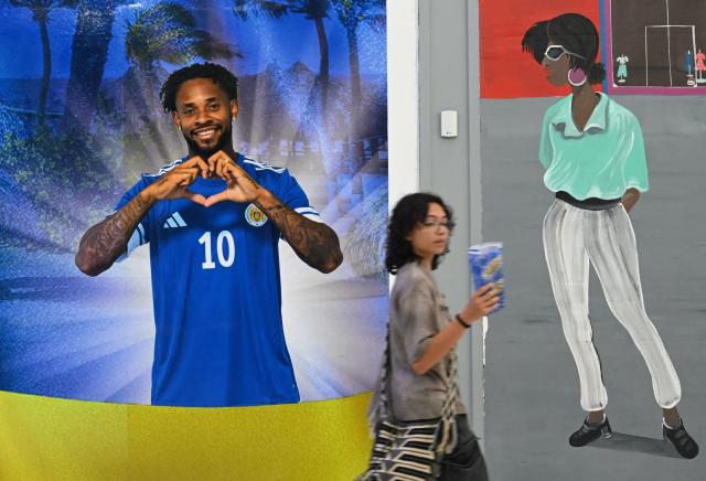 A woman walks an image of Curacao's player Leandro Bacuna at the official fan store in Willemstad, Curacao, in the Dutch Caribbean, on April 8, 2026. Small island, big dreams. The poster of Curacao’s national football team hangs at the entrance of Marchena, a working-class neighborhood of Willemstad, the capital of the smallest territory ever to qualify for a World Cup. (Photo by Raul ARBOLEDA / AFP)