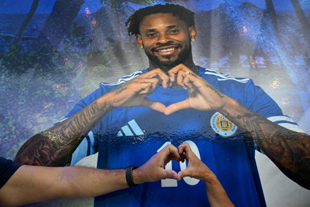 A couple makes a heart next to an image of Curacao's player Leandro Bacuna at the official fan store in Willemstad, Curacao, in the Dutch Caribbean, on April 8, 2026. Small island, big dreams. The poster of Curacao’s national football team hangs at the entrance of Marchena, a working-class neighborhood of Willemstad, the capital of the smallest territory ever to qualify for a World Cup. (Photo by Raul ARBOLEDA / AFP)