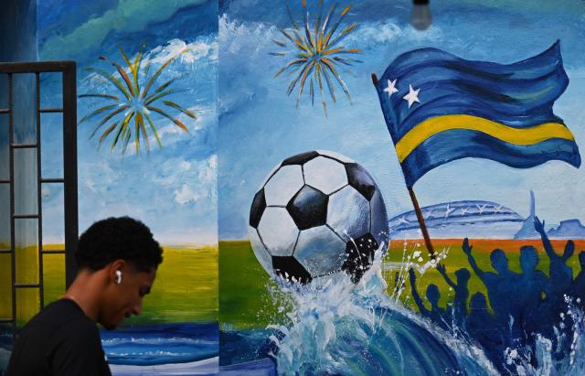 A man walks past a mural depicting a football and a flag of Curacao in Willemstad, in the Dutch Caribbean, on April 10, 2026. Small island, big dreams. The poster of Curacao’s national football team hangs at the entrance of Marchena, a working-class neighborhood of Willemstad, the capital of the smallest territory ever to qualify for a World Cup. (Photo by Raul ARBOLEDA / AFP)