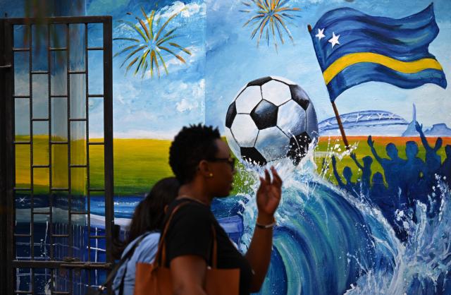 Women walk past a mural depicting a football and a flag of Curacao in Willemstad, in the Dutch Caribbean, on April 10, 2026. Small island, big dreams. The poster of Curacao’s national football team hangs at the entrance of Marchena, a working-class neighborhood of Willemstad, the capital of the smallest territory ever to qualify for a World Cup. (Photo by Raul ARBOLEDA / AFP)