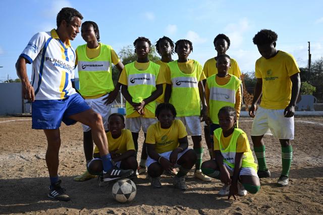 Dutch-Curacaoan former football player and manager Remko Bicentini (L) poses with his players as part of their training session at the Bicentini Foundation football school in the Fuik neighborhood, east of Willemstad, in the Dutch Caribbean, on April 11, 2026. Small island, big dreams. The poster of Curacao’s national football team hangs at the entrance of Marchena, a working-class neighborhood of Willemstad, the capital of the smallest territory ever to qualify for a World Cup. (Photo by Raul ARBOLEDA / AFP)