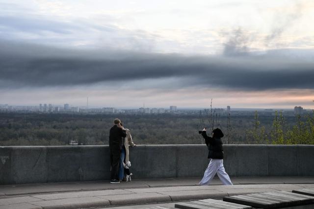 People pose for pictures as smoke rises over Kyiv following a Russian strike on April 16, 2026, amid the Russian invasion of Ukraine. Russian strikes killed at least 12 people in Ukraine, local authorities said on April 16, after Moscow pummelled its neighbour in overnight attacks. (Photo by Genya SAVILOV / AFP)