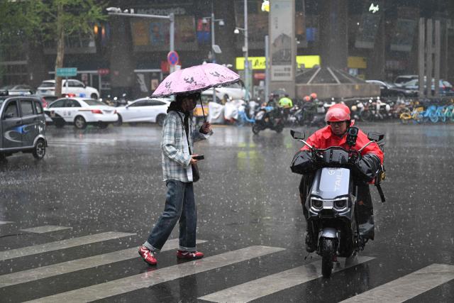A man rides his scooter past a woman holding an umbrella as she crosses a street during a rainy day in Beijing on April 16, 2026. (Photo by Pedro PARDO / AFP)