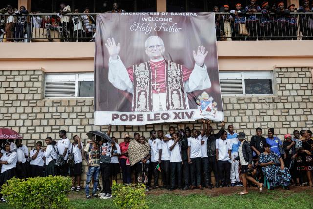Attendees use a banner displaying a picture of Pope Leo XIV on to shelter from the rain as they wait for him to arrive to lead the Holy Mass at Bamenda Airport in Bamenda, on the fourth day of an 11-day apostolic journey to Africa, on April 16, 2026. (Photo by Patrick MEINHARDT / AFP)