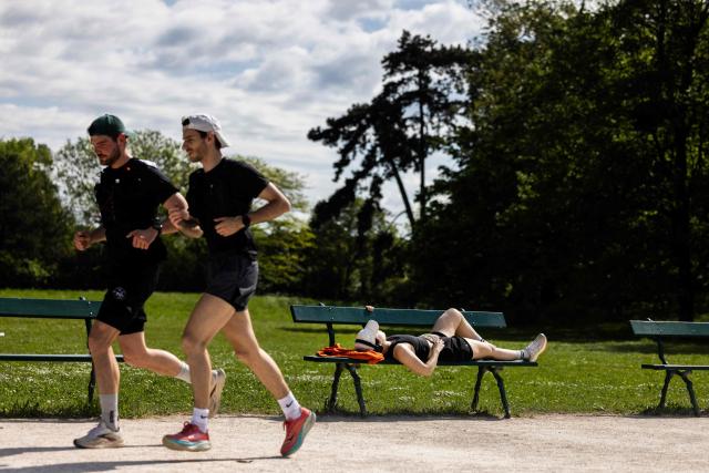 Two men run past a woman lying on a bench at the Bois de Vincennes park in Paris on April 18, 2026. (Photo by Behrouz MEHRI / AFP)