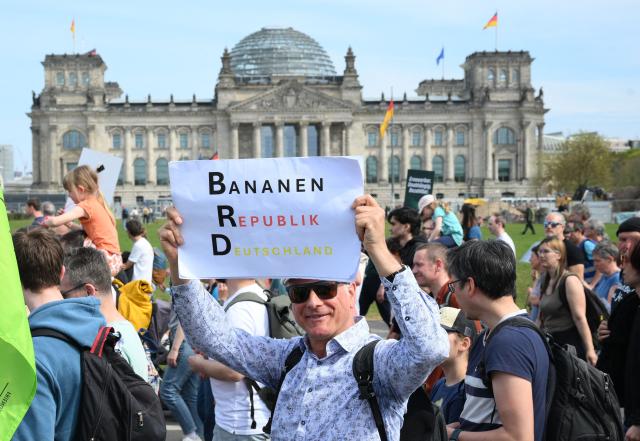 A protester walking past the Reichstag building that houses the Bundestag (lower house of parliament) holds up a placard reading "Banana Republic Germany" during a demonstration of activists and supporters of Fridays for Future, Campact and others against the German government's energy policy, on April 18, 2026 in Berlin. Activists call for faster expansion of solar and wind energy, rapid development of distribution grids, storage and digitalization, a strong European clean energy industry, lower electricity taxes and cheaper power for heat pumps, as well as social support and tenant protection to ensure affordable, climate-friendly heating. (Photo by RALF HIRSCHBERGER / AFP)