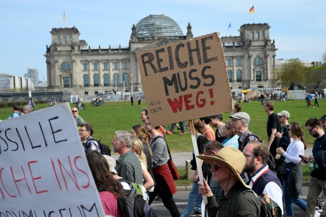 A protester walking past the Reichstag building that houses the Bundestag (lower house of parliament) holds up a placard reading "Reiche muss weg!" (Reiche must go, alluding to German Economy and Energy Minister Katherina Reiche) during a demonstration of activists and supporters of Fridays for Future, Campact and others against the German government's energy policy, on April 18, 2026 in Berlin. Activists call for faster expansion of solar and wind energy, rapid development of distribution grids, storage and digitalization, a strong European clean energy industry, lower electricity taxes and cheaper power for heat pumps, as well as social support and tenant protection to ensure affordable, climate-friendly heating. (Photo by RALF HIRSCHBERGER / AFP)