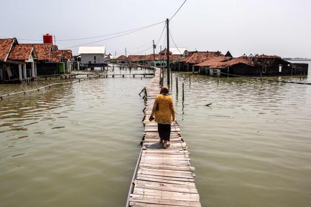 A woman walks across a bridge past houses partially submerged in seawater due to tidal flooding at the Timbulsloko village in Demak, Central Java on April 18, 2026. (Photo by DEVI RAHMAN / AFP)