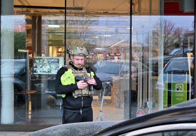 A policeman looks at his phone in front of a hole made from a bullet in the window of a supermarket after a shooting in Kyiv on April 18, 2026. A gunman opened fire in Kyiv on April 18, 2026, killing at least five people, wounding others and taking hostages at a supermarket before being killed during an arrest attempt, officials said. Ukrainian President said that at least five people were dead and 10 hospitalised with wounds and trauma after the shooting in a residential district of the capital. (Photo by Sergei SUPINSKY / AFP)