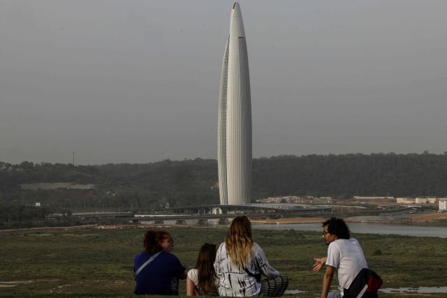 People sit in a park looking towards the Mohammed VI Tower, approximately 250 meters tall with nearly 55 floors, the tallest tower in Morocco, comprising a hotel, apartments, and restaurants, in Sale on April 20, 2026. (Photo by Abdel Majid BZIOUAT / AFP)