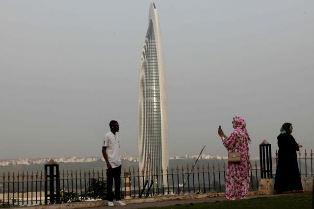 Tourists pose for a photograph with the tallest tower in Morocco, the Mohammed VI Tower in the background, comprising of a hotel, apartments, and restaurants, in Sale on April 20, 2026. (Photo by Abdel Majid BZIOUAT / AFP)