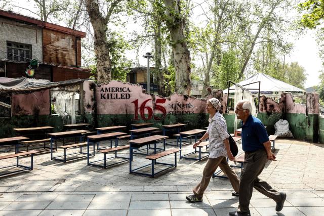 An Iranian couple walks past symbolic wall art that reads, “America killed 165 Minab's Students,” referring to an elementary school in Minab, southern Iran, which was allegedly hit by a US-Israeli missile on the first day of the war, killing over 100 schoolchildren, in northern Tehran on April 21, 2026, amid a ceasefire in the region. With the end of a two-week ceasefire approaching, the White House said US Vice President was ready to return to Pakistan for fresh negotiations to end a conflict that has sent crude soaring and revived inflation fears. (Photo by AFP) / 