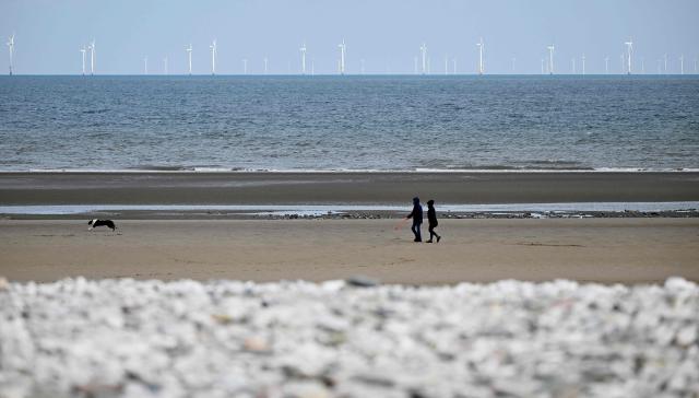 The Ryhl Flats windfarm is pictured on the horizon in the Irish Sea as dog walkers stroll on the beach at Abergele in north Wales on April 21, 2026. Britain's government on April 21, 2026, unveiled plans to accelerate its clean energy drive after oil and gas prices soared in the wake of the US-Iran war. Placing solar panels and wind turbines on industrial and railway sites could power about five million homes, the Labour government said in a statement. (Photo by Paul ELLIS / AFP)