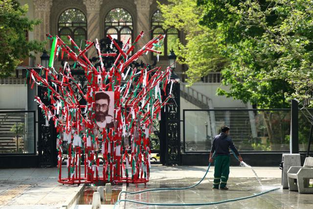 An Iranian municipal worker sprays water on the walkway of a park near a structure adorned with the national flag and a portrait of the late Iranian supreme leader Ayatollah Ali Khamenei as a young man,  in northern Tehran on April 21, 2026, amid a ceasefire in the region. With the end of a two-week ceasefire approaching, the White House said US Vice President was ready to return to Pakistan for fresh negotiations to end a conflict that has sent crude soaring and revived inflation fears. (Photo by AFP) / 
