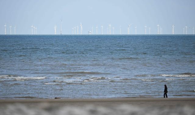 The Ryhl Flats windfarm is pictured on the horizon in the Irish Sea as a dog walker strolls on the beach at Abergele in north Wales on April 21, 2026. Britain's government on April 21, 2026, unveiled plans to accelerate its clean energy drive after oil and gas prices soared in the wake of the US-Iran war. Placing solar panels and wind turbines on industrial and railway sites could power about five million homes, the Labour government said in a statement. (Photo by Paul ELLIS / AFP)
