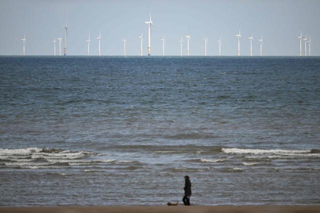 The Ryhl Flats windfarm is pictured on the horizon in the Irish Sea as a dog walker strolls on the beach at Abergele in north Wales on April 21, 2026. Britain's government on April 21, 2026, unveiled plans to accelerate its clean energy drive after oil and gas prices soared in the wake of the US-Iran war. Placing solar panels and wind turbines on industrial and railway sites could power about five million homes, the Labour government said in a statement. (Photo by Paul ELLIS / AFP)