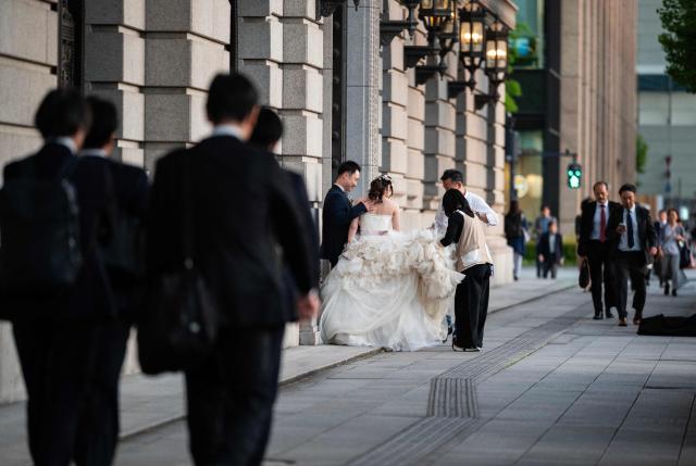 Businessmen walk past a couple having an evening photo shoot in their wedding outfits in Tokyo on April 21, 2026, near the Tokyo Station complex, one of a number of popular photo spots for newlyweds. (Photo by Andrew CABALLERO-REYNOLDS / AFP)
