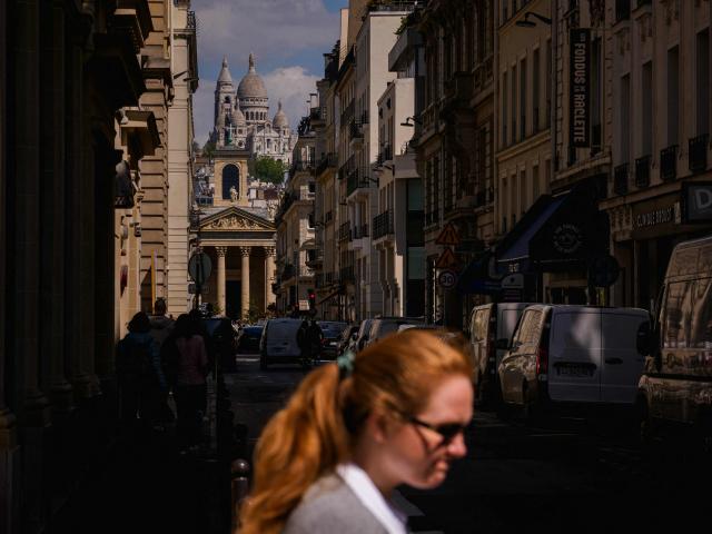 A woman walks down a street with the Sacre-Coeur Basilica on the Montmartre hill in the background, in Paris on April 21, 2026. (Photo by Dimitar DILKOFF / AFP)
