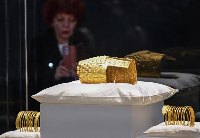 A guest takes photos of the ancient Cotofenesti helmet and two Dacian gold bracelets on display at the National Museum of History of Romania (MNIR) in Bucharest on April 21, 2026. The artifacts, shown together for the first time since their repatriation, are part of a national campaign to bring Romania’s most significant ancient treasures to the public across the country. In January 2025, the Cotofenesti helmet and several gold bracelets were stolen from the Drents Museum in Assen, Netherlands. They were on loan from the National Museum of History of Romania (MNIR) as part of the "Dacia – Empire of Gold and Silver" exhibition. (Photo by Daniel MIHAILESCU / AFP)