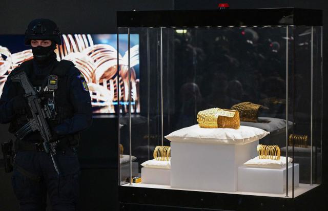 An armed police officer stands next to the ancient Cotofenesti helmet and two Dacian gold bracelets on display at the National Museum of History of Romania (MNIR) in Bucharest on April 21, 2026. The artifacts, shown together for the first time since their repatriation, are part of a national campaign to bring Romania’s most significant ancient treasures to the public across the country. In January 2025, the Cotofenesti helmet and several gold bracelets were stolen from the Drents Museum in Assen, Netherlands. They were on loan from the National Museum of History of Romania (MNIR) as part of the "Dacia – Empire of Gold and Silver" exhibition. (Photo by Daniel MIHAILESCU / AFP)