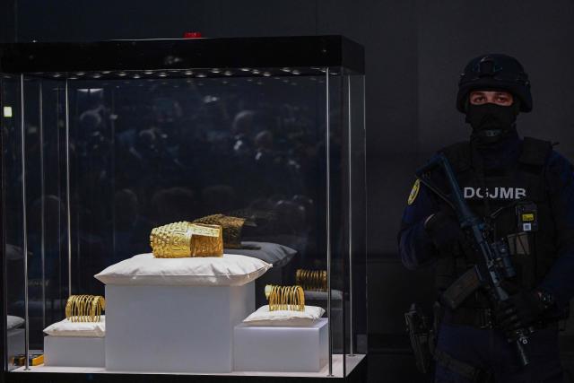 An armed police officer stands next to the ancient Cotofenesti helmet and two Dacian gold bracelets on display at the National Museum of History of Romania (MNIR) in Bucharest on April 21, 2026. The artifacts, shown together for the first time since their repatriation, are part of a national campaign to bring Romania’s most significant ancient treasures to the public across the country. In January 2025, the Cotofenesti helmet and several gold bracelets were stolen from the Drents Museum in Assen, Netherlands. They were on loan from the National Museum of History of Romania (MNIR) as part of the "Dacia – Empire of Gold and Silver" exhibition. (Photo by Daniel MIHAILESCU / AFP)