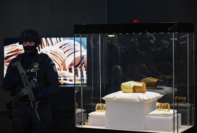An armed police officer stands next to the ancient Cotofenesti helmet and two Dacian gold bracelets on display at the National Museum of History of Romania (MNIR) in Bucharest on April 21, 2026. The artifacts, shown together for the first time since their repatriation, are part of a national campaign to bring Romania’s most significant ancient treasures to the public across the country. In January 2025, the Cotofenesti helmet and several gold bracelets were stolen from the Drents Museum in Assen, Netherlands. They were on loan from the National Museum of History of Romania (MNIR) as part of the "Dacia – Empire of Gold and Silver" exhibition. (Photo by Daniel MIHAILESCU / AFP)