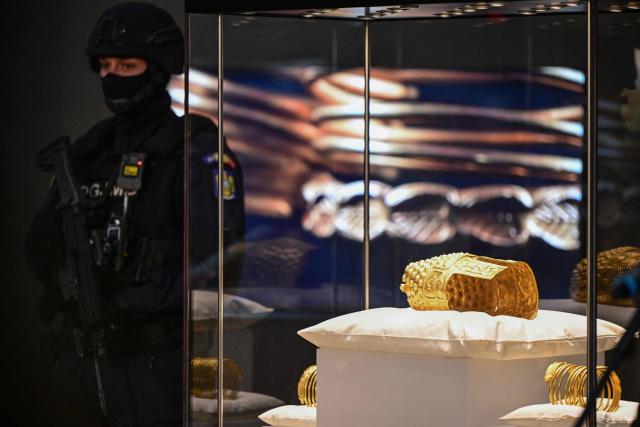 An armed police officer stands next to the ancient Cotofenesti helmet and two Dacian gold bracelets on display at the National Museum of History of Romania (MNIR) in Bucharest on April 21, 2026. The artifacts, shown together for the first time since their repatriation, are part of a national campaign to bring Romania’s most significant ancient treasures to the public across the country. In January 2025, the Cotofenesti helmet and several gold bracelets were stolen from the Drents Museum in Assen, Netherlands. They were on loan from the National Museum of History of Romania (MNIR) as part of the "Dacia – Empire of Gold and Silver" exhibition. (Photo by Daniel MIHAILESCU / AFP)