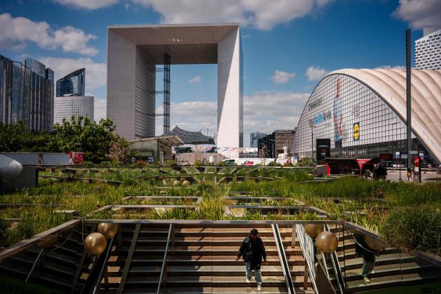 A woman walks down the stairs with a background view of The Great Arch of the Defense in the La Defense business district in Puteaux outside Paris on April 21, 2026. (Photo by Dimitar DILKOFF / AFP)