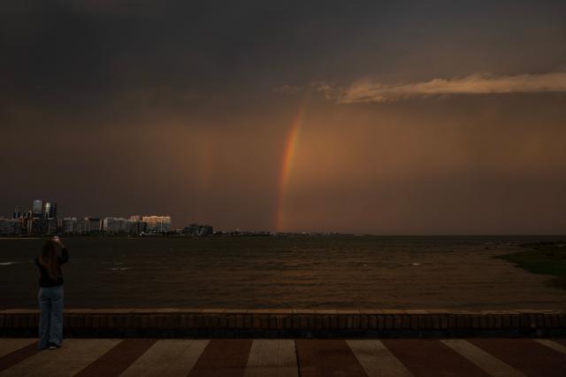 A woman takes a picture of a rainbow as the sun sets after rainfall in Montevideo on April 21, 2026. (Photo by Eitan ABRAMOVICH / AFP)