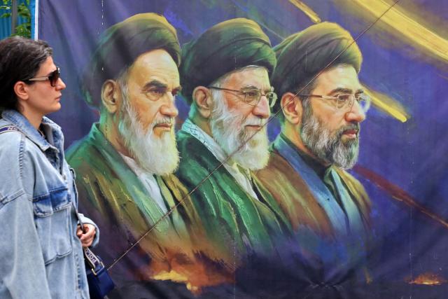 This photo taken on April 19, 2026 shows a woman walking past a banner featuring Iran's late supreme leaders Ayatollah Ruhollah Khomeini (L) and Ayatollah Ali Khamenei (C) next to newly elected supreme leader Ayatollah Mojtaba Khamenei, in Tehran. Since the outbreak of war between Iran, Israel and the United States, colourful murals have sprung up across Tehran and beyond, rich in symbolism and themes of resistance and defiance. Murals and banners have long been a defining feature of Iran's urban landscape, particularly in Tehran, where such paintings in central squares often mirror the state's political messaging and foreign policy stance. (Photo by ATTA KENARE / AFP) / Attention editors: AFP covers the war in the Middle East through its extensive regional network, including bureaus in Tehran, Jerusalem and several neighboring countries.Since the start of the conflict, journalists have been working under increasingly restrictive conditions. Authorities in several countries have limited reporters movements, photo and live video coverage from sensitive locations. Some governments and armed groups have banned images of missile or drone strikes and other security-related sites. / 