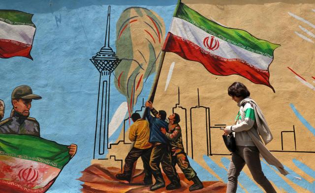 This photo taken on April 21, 2026 shows an Iranian woman walking past an anti-USA and anti-Israel mural, in Tehran, amid a ceasefire in the region. Since the outbreak of war between Iran, Israel and the United States, colourful murals have sprung up across Tehran and beyond, rich in symbolism and themes of resistance and defiance. Murals and banners have long been a defining feature of Iran's urban landscape, particularly in Tehran, where such paintings in central squares often mirror the state's political messaging and foreign policy stance. (Photo by ATTA KENARE / AFP) / Attention editors: AFP covers the war in the Middle East through its extensive regional network, including bureaus in Tehran, Jerusalem and several neighboring countries.Since the start of the conflict, journalists have been working under increasingly restrictive conditions. Authorities in several countries have limited reporters movements, photo and live video coverage from sensitive locations. Some governments and armed groups have banned images of missile or drone strikes and other security-related sites. / 
