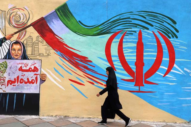 This photo taken on April 21, 2026 shows an Iranian woman walking past an anti-USA and anti-Israel mural, in Tehran, amid a ceasefire in the region. Since the outbreak of war between Iran, Israel and the United States, colourful murals have sprung up across Tehran and beyond, rich in symbolism and themes of resistance and defiance. Murals and banners have long been a defining feature of Iran's urban landscape, particularly in Tehran, where such paintings in central squares often mirror the state's political messaging and foreign policy stance. (Photo by ATTA KENARE / AFP) / Attention editors: AFP covers the war in the Middle East through its extensive regional network, including bureaus in Tehran, Jerusalem and several neighboring countries.Since the start of the conflict, journalists have been working under increasingly restrictive conditions. Authorities in several countries have limited reporters movements, photo and live video coverage from sensitive locations. Some governments and armed groups have banned images of missile or drone strikes and other security-related sites. / 