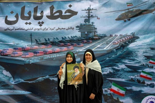 This photo taken on February 11, 2026 shows women posing with a poster depicting Iran's supreme leader Ayatollah Ali Khamenei as they stand before a banner depicting a US Navy aircraft carrier with US-flag-wrapped coffins on the deck, during a rally marking the 47th anniversary of the 1979 Islamic revolution in Tehran. Murals and banners have long been a defining feature of Iran's urban landscape, particularly in Tehran, where such paintings in central squares often mirror the state's political messaging and foreign policy stance. (Photo by AFP) / Attention editors: AFP covers the war in the Middle East through its extensive regional network, including bureaus in Tehran, Jerusalem and several neighboring countries.Since the start of the conflict, journalists have been working under increasingly restrictive conditions. Authorities in several countries have limited reporters movements, photo and live video coverage from sensitive locations. Some governments and armed groups have banned images of missile or drone strikes and other security-related sites. / 