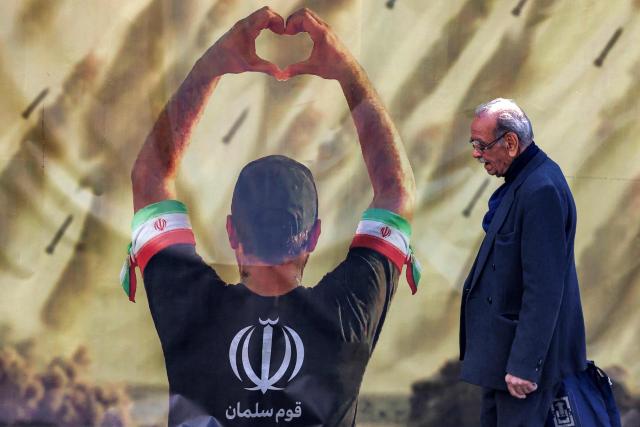 This photo taken on April 6, 2026 shows a man walking past a banner depicting a man holding up a heart gestures before missiles being fired, in Tehran. Since the outbreak of war between Iran, Israel and the United States, colourful murals have sprung up across Tehran and beyond, rich in symbolism and themes of resistance and defiance. Murals and banners have long been a defining feature of Iran's urban landscape, particularly in Tehran, where such paintings in central squares often mirror the state's political messaging and foreign policy stance. (Photo by ATTA KENARE / AFP) / 