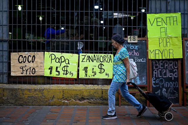 A woman pulls a shopping bag with wheels past a supermarket displaying signs with food prices in US dollars in Caracas on April 22, 2026. (Photo by Federico PARRA / AFP)