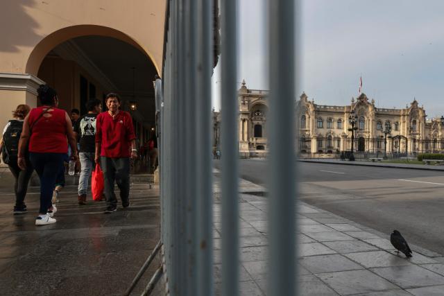 People walk along the perimeter of Plaza Mayor, behind metal fences that keep the square closed, with the Government Palace in the background, in Lima on April 22, 2026. Peru's foreign and defense ministers resigned on April 22 in protest over the interim president's decision to stall a $3.5 billion deal for the purchase of 24 US F-16 fighter jets. (Photo by Connie FRANCE / AFP)