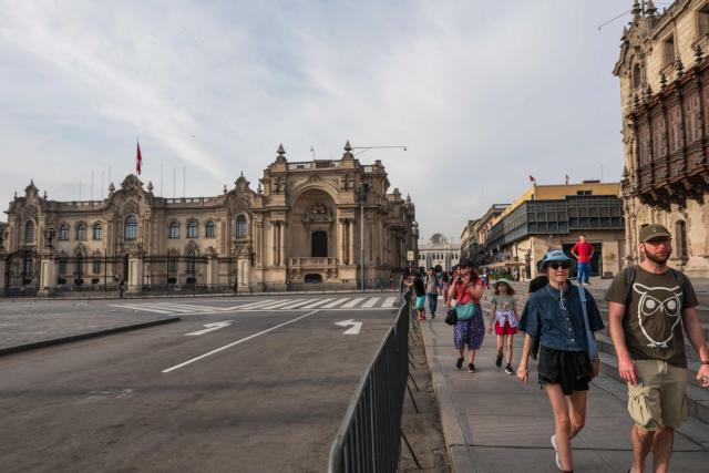 Tourists walk along the perimeter of Plaza Mayor, behind metal fences that keep the square closed, with the Government Palace in the background, in Lima on April 22, 2026. Peru's foreign and defense ministers resigned on April 22 in protest over the interim president's decision to stall a $3.5 billion deal for the purchase of 24 US F-16 fighter jets. (Photo by Connie FRANCE / AFP)