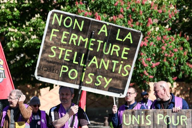 An employee of multinational automotive manufacturing corporation Stellantis holds a placard reading "No to the closure of Stellantis Poissy" during a demonstration called by trade unions outside the Poissy City Hall, in the outskirts of Paris, on April 23, 2026. The car manufacturer Stellantis announced on April 16, 2026 that it would cease car production at its historic Poissy plant after 2028, thereby turning a page in the industrial history of the Paris region. (Photo by ALAIN JOCARD / AFP)