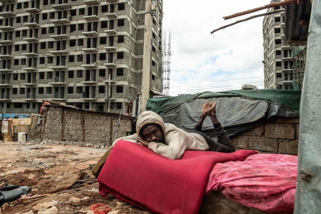 Samson Kiongo, 40, a resident of Soweto Zone D, rests on a mattress amid the remains of his demolished house in front of buildings under construction for a state affordable housing programme following government-planned demolitions carried out to make space for the project at the Kibera informal settlement, in Nairobi on April 23, 2026. Kenyas Affordable Housing Programme is a government initiative to replace informal settlements with low-cost housing. In Kibera, the largest informal settlement in Kenya, demolitions have cleared some areas for new developments aimed at improving living conditions and home ownership. However, the programme has faced controversy over evictions, compensation and displacement of residents before completion of new housing. (Photo by Gordwin Odhiambo / AFP)