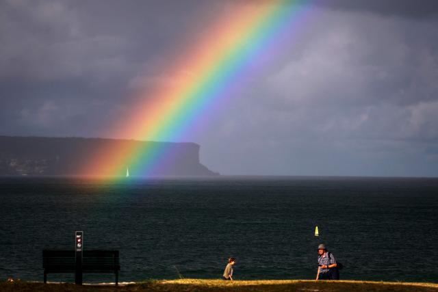 A sailing boat passes a rainbow as people walk along Balmoral Beach on Sydney Harbour on April 24, 2026. (Photo by DAVID GRAY / AFP)