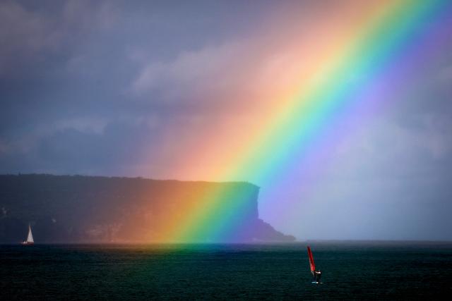 A rainbow can be seen near a sailing boat and a man riding a sailboard in front of North Head on Sydney Harbour on April 24, 2026. (Photo by DAVID GRAY / AFP)