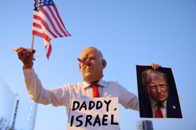 TOPSHOT - An Israeli left-wing activist, disguised as the Israeli Prime Minister Benjamin Netanyahu and holding an image of the US President Donald Trump and the US flag, demonstrates in HaBima Square against the ongoing war with Iran and against the Israeli government, in Tel Aviv on April 25, 2026. Since the ceasefire between Israel, the United States, and Iran came into effect, a relative calm has prevailed, with no bombings reported in Iran or the Gulf, after five weeks of conflict that left thousands dead, primarily in Iran and Lebanon. (Photo by Jack GUEZ / AFP) / 