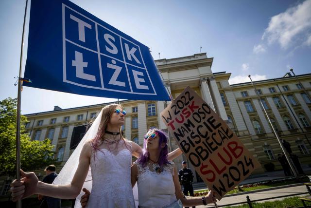 A couple holds banners reading 'Tusk is lying' and 'In 2022, Tusk personally promised us a wedding in 2024' as they take part in a demonstration of LGBT organisations in front of Poland's Prime Minister Chancellery building in Warsaw on April 25, 2026. The LGBT organisations point out that the Polish government has not implemented landmark rulings of the European Court of Human Rights that require Poland to recognize same-sex marriages concluded in other EU member states. (Photo by Wojtek RADWANSKI / AFP)