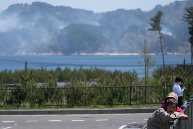 Two women watch a fire burn as smoke rises from a hillside in the background in the town of Otsuchi in Iwate Prefecture on April 26, 2026. Hundreds of firefighters were battling wildfires in the forests of northern Japan, as authorities urged more than 3,200 people to evacuate from their homes, government officials said. (Photo by ANDREW CABALLERO-REYNOLDS / AFP)