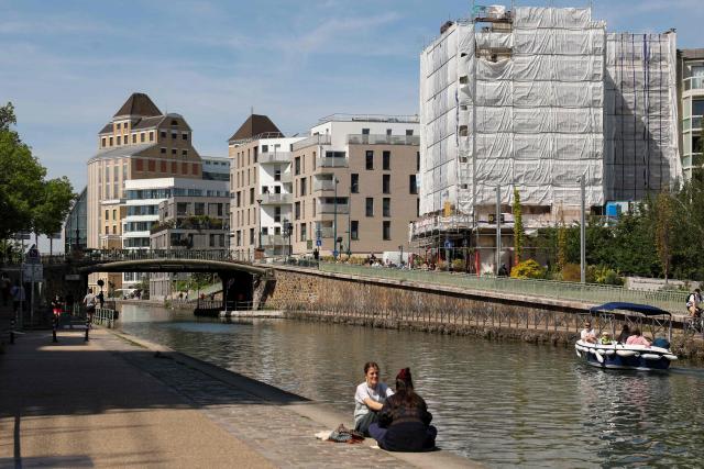 People sit by the Canal de l'Ourcq with the Grands Moulins de Pantin in the background in Pantin, suburb of Paris on April 26, 2026. (Photo by Charlotte SIEMON / AFP)