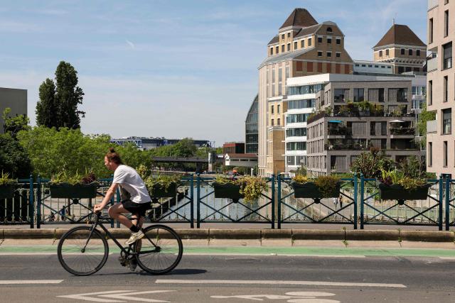 A cyclist rides his bike on a bridge crossing the Canal de l'Ourcq with the Grands Moulins de Pantin in the background in Pantin, suburb of Paris on April 26, 2026. (Photo by Charlotte SIEMON / AFP)